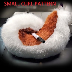 Może przedstawiać: Biało-brązowy ogon z futra syntetycznego z małym lokiem na końcu. Ogon leży na czarnej powierzchni z maszyną do szycia w tle.