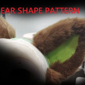Puede incluir: Primer plano de una oreja peluda marrón con un forro interior de tela verde. La oreja está fijada a una superficie de espuma blanca. El texto "EAR SHAPE PATTERN" es visible en la parte superior de la imagen.