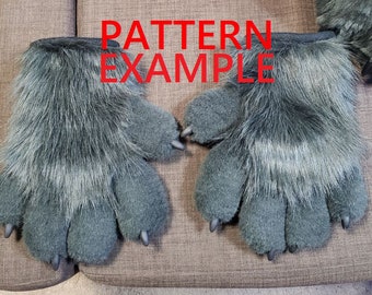 May include: A pair of grey furry hand paws with black claws. The paws are laid out flat on a brown fabric surface. The text "PATTERN EXAMPLE" is written in red letters above the paws.