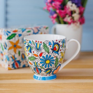 May include: A white ceramic mug with a colorful floral pattern. The mug has a blue rim and a handle. The mug is sitting on a wooden surface in front of a vase of flowers.