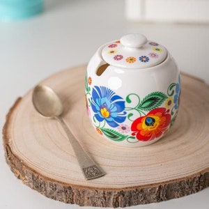 May include: White ceramic sugar bowl with a floral design in shades of blue, red, green, and yellow. The bowl has a lid and a spoon rest. The bowl is sitting on a wooden slice.