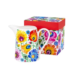 Op de afbeelding: Witte keramische kan met een levendig bloemenpatroon in geel, rood, blauw en paars. De kan heeft een gebogen handvat en tuit. Het staat naast een bijpassende geschenkdoos met een rode bovenkant en bloemenpatroon.