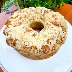 May include: A round cake with a crumb topping. The cake is light brown and has a hole in the center. It is sitting on a white cake stand.