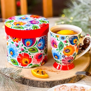 May include: A white ceramic mug with a colorful floral pattern and a red rim, filled with a hot beverage and a slice of lemon. The mug is sitting on a wooden surface next to a white box with a similar floral pattern and a red lid.