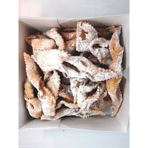 Pu&ograve; includere: Una scatola piena di pasticcini dorati e fritti, generosamente cosparsi di zucchero a velo. I pasticcini hanno una forma attorcigliata a nastro e sono disposti in una scatola di cartone bianca. Vista dall'alto.