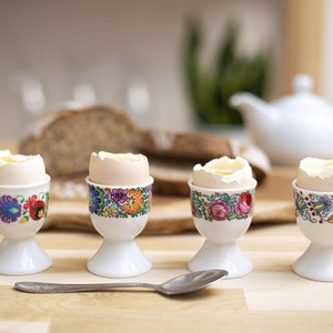 Può includere: Quattro porta uova in ceramica bianca con motivi floreali colorati contengono uova sode. I porta uova sono su un tavolo di legno con un cucchiaio e una pagnotta di pane sullo sfondo.