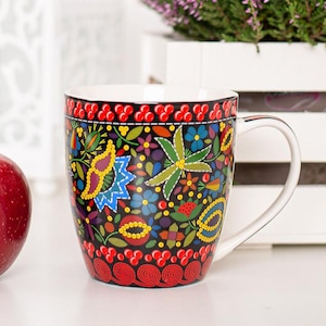 May include: A white ceramic mug with a black background and colorful floral pattern. The pattern features red, yellow, blue, green, and purple flowers and leaves. The mug has a white handle.