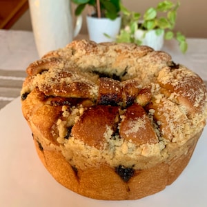May include: A close-up of a round, golden brown bread with a crumb topping. The bread has a slightly sweet, caramelized glaze and is baked in a ring shape.