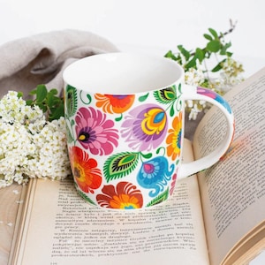 Può includere: Una tazza in ceramica bianca con un motivo floreale colorato. La tazza è appoggiata su un libro con una copertina marrone.