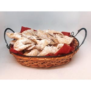 Pu&ograve; includere: Un cestino di vimini con fodera in tessuto rosso, pieno di pasticcini fritti spolverati di zucchero a velo. Il cestino ha un manico in metallo scuro e si trova su uno sfondo bianco. I pasticcini sono dorati e croccanti.