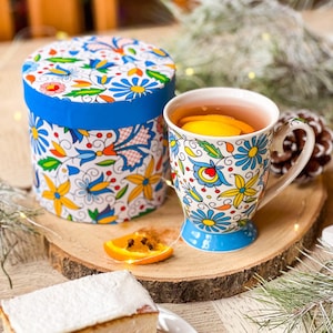 May include: A white mug with a blue rim and colorful floral pattern, filled with tea and a slice of lemon, sitting on a wooden coaster next to a gift box with a similar floral pattern.