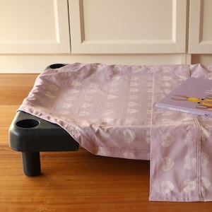 Op de afbeelding: Een zwart huisdierenbed met een lavendelkleurig laken met witte hartjes. Een kinderboek getiteld "Llama Llama Red Pajama" ligt op het laken. Het bed staat op een houten vloer.