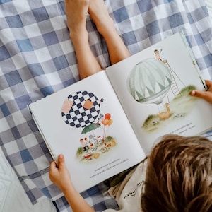 Puede incluir: Las manos de un niño sostienen un libro infantil colorido abierto con ilustraciones de globos aerostáticos, un cisne y otros objetos caprichosos. El libro está abierto sobre una manta a cuadros azul y blanco.