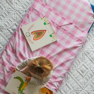 May include: A young child sits on a pink and white gingham blanket reading a book about animals. The book has illustrations of a crocodile and a koala. The child is wearing a white shirt and has their hair tied back in a ponytail.