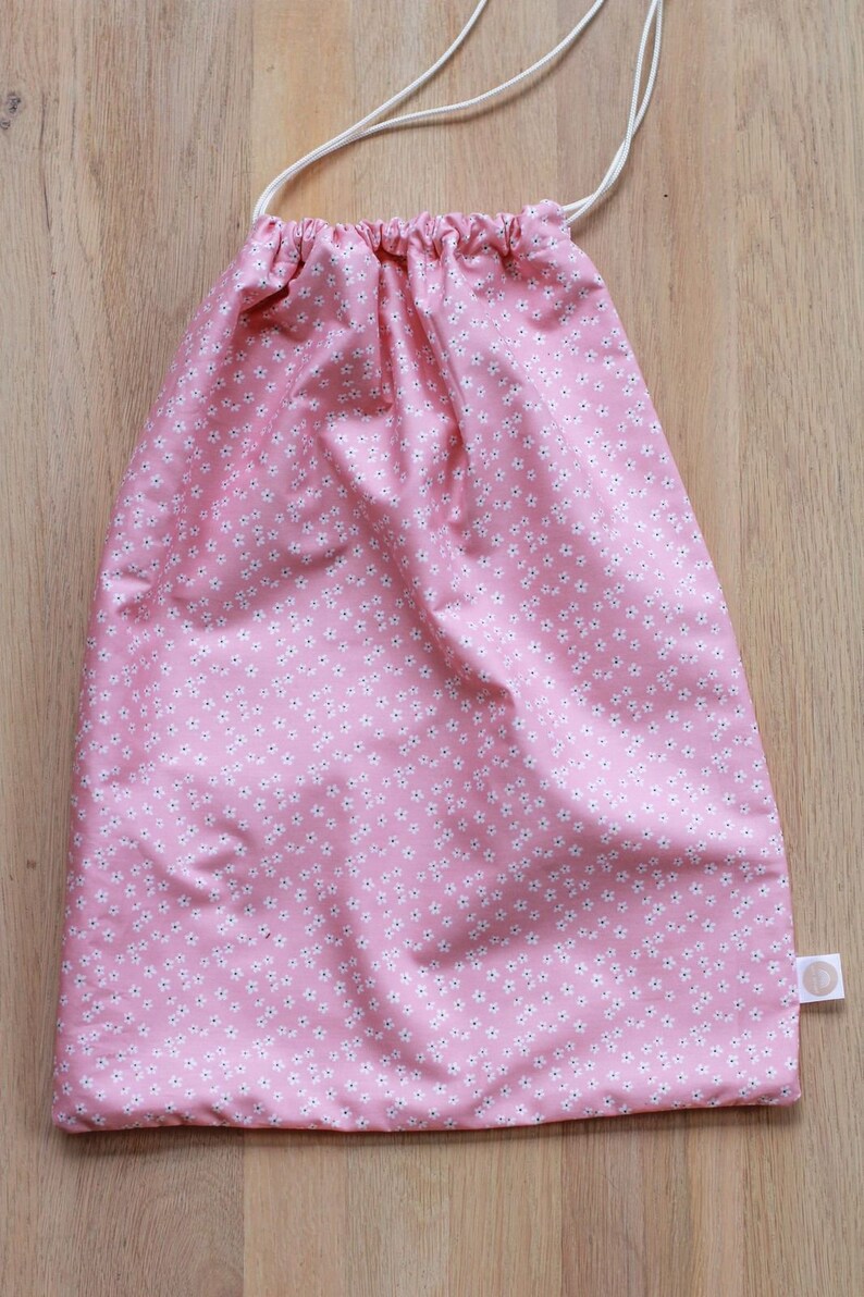 K&ouml;nnte beinhalten: Eine rosa Kordeltasche mit einem wei&szlig;en Blumenmuster. Die Tasche hat eine wei&szlig;e Kordel und ein kleines wei&szlig;es Etikett.