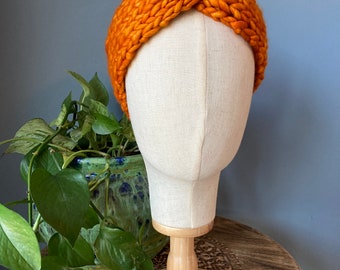 May include: A bright orange knitted headband with a knotted design. The headband is displayed on a fabric-covered mannequin head.