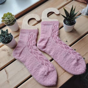 May include: A pair of light pink knitted socks with a decorative cable pattern. The socks are displayed on wooden hangers, with small potted plants in the background. The socks are made of a soft, textured yarn.