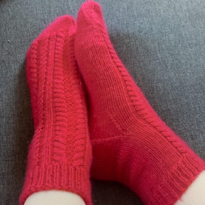 Puede incluir: Un par de calcetines de punto rojo brillante con un diseño de punto trenzado en la parte delantera y un puño acanalado. Los calcetines están hechos de un hilo suave y texturizado, presentados sobre una superficie gris.