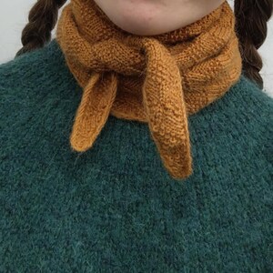 May include: A close-up of a mustard-colored knitted scarf tied around the neck. The scarf is made of a textured yarn and is knotted in the front. The scarf is worn over a teal-colored sweater.