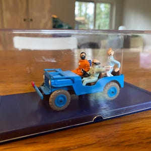 Peut inclure: Un modèle réduit de jeep bleue avec trois figurines à l'intérieur, présenté dans un boîtier en plastique transparent. La jeep a un pare-chocs avant rouge et des roues bleues. Les figurines portent des tenues orange, bleu et beige.