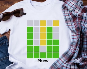 Peut inclure: T-shirt blanc avec un motif en grille vert, gris et jaune et le mot "Phew" imprimé en dessous. Le t-shirt est posé sur une surface en bois avec des lunettes de soleil, un jean bleu et une chemise à carreaux.