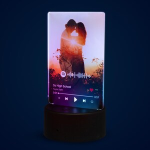 May include: A rectangular, illuminated night light featuring a romantic image of a couple embracing at sunset. The night light displays a music player interface with the song "So High School" by Taylor Swift. The base is a dark, rounded square.