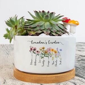 May include: White ceramic planter with a wooden base, featuring the text "Grandma's Garden" and a row of colourful flowers with names written below each flower. The planter is filled with green succulents and a small red and yellow mushroom.