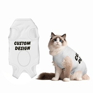 May include: White pet tank top with the text "CUSTOM DESIGN" in black. A fluffy cat wearing a similar tank top is also in the image. The cat has blue eyes and is sitting on a white background. Pet apparel.