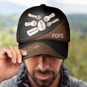 Puede incluir: Gorra de béisbol negra y marrón con la palabra "POPS" y nombres como "Sebastian" y "Monica" impresos. La gorra presenta un diseño de corazón y latido del corazón, junto con ilustraciones de puños y huellas de patas.