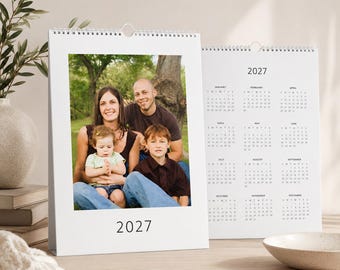 May include: A white wall calendar for the year 2027 featuring a spiral binding at the top. The front page displays a color photograph of a family of three, including two adults and a young child, with the year 2027 printed in black at the bottom.