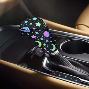 May include: A black gear shift cover adorned with a celestial pattern of stars, moons, and planets in shades of blue, green, purple, and pink. The cover is installed in a car, with the gear shift in the center console.