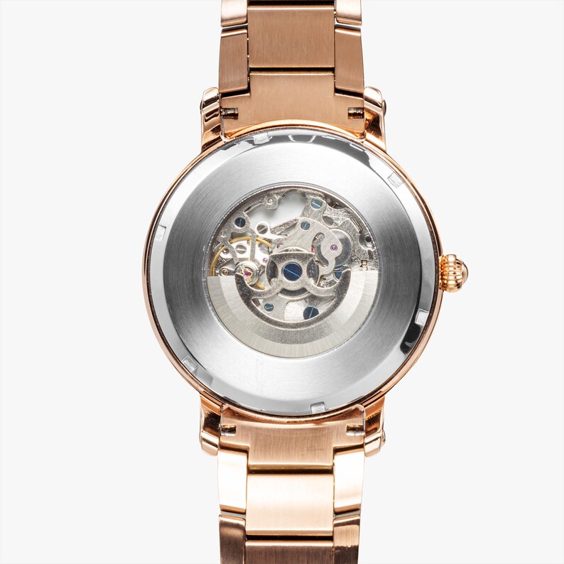 May include: Rose gold-toned wristwatch with a visible mechanical movement. The watch features a round face with a clear back, showcasing the intricate inner workings. The band is made of matching rose gold links.
