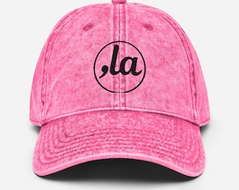 Peut inclure: Casquette de baseball rose avec un style vintage. La casquette a une visière incurvée et une couronne arrondie. Un graphisme circulaire noir avec les lettres ",la" est centré sur le devant. La casquette a un aspect légèrement délavé.