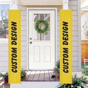 May include: Two vertical yellow banners hanging on either side of a white front door. Each banner features the text CUSTOM DESIGN printed in bold black capital letters. The door has a glass window with a green wreath hanging in the center.