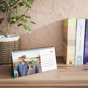 Puede incluir: Un calendario de escritorio para mayo con una foto familiar, encuadernado en espiral en la parte superior. El calendario está sobre una superficie de madera, con libros y una planta en maceta en el fondo. El calendario muestra los días del mes.