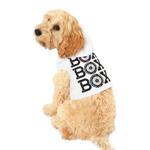 Op de afbeelding: Een witte hondenhoodie met een zwarte afbeelding van het woord "BOX" dat wordt herhaald met bandenprofielpatronen. De hoodie is op een lichtbruine hond.