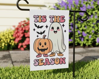 May include: A white Halloween garden flag featuring a cartoon ghost and pumpkin design. The flag reads "TIS THE SEASON" in pink and orange lettering. Black bats and stars are also featured in the design. The flag is displayed on a black metal stand in a grassy garden.