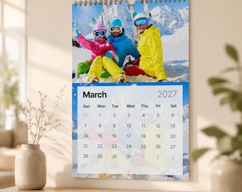 Könnte beinhalten: Ein Wandkalender für März 2027 mit einem Foto von drei Personen in Skiausrüstung, darunter eine rosa, eine blaue und eine gelbe Jacke, in einer verschneiten Berglandschaft. Das Kalenderraster zeigt die Tage des Monats an.