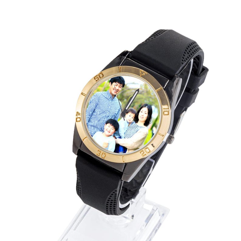 May include: A black wristwatch with a gold-coloured bezel and a black rubber strap. The watch face displays a family portrait. The watch is displayed on a clear stand.