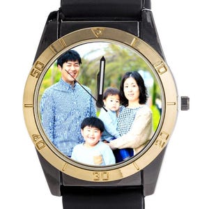 May include: Black wristwatch with a gold-coloured bezel and a black strap. The watch face displays a family portrait. The watch has a black dial with white numerals and hands.