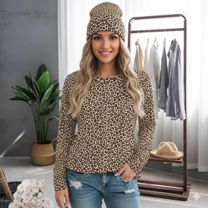 Puede incluir: Una mujer con un gorro y una camisa de manga larga con estampado de leopardo a juego. La camisa y el gorro presentan un estampado de manchas marrones y beige. Lleva vaqueros azules. Un perchero con varias prendas en el fondo.