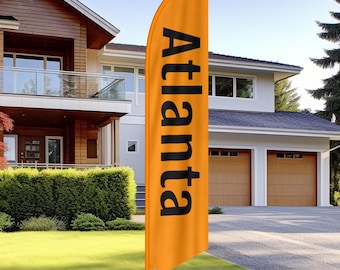 May include: An orange feather flag with the word "Atlanta" printed in black. The flag is displayed outdoors in front of a house with a green lawn and a driveway.