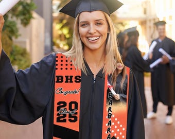 May include: A person wearing a black graduation gown and cap with an orange and black graduation sash. The sash has the text "BHS Congrats! Class of 2025" and a photo of a person. The sash also has the name "Barbara" written vertically.