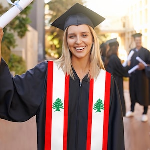 Puede incluir: Una graduada con birrete y toga negra, sosteniendo un diploma. La toga presenta una banda blanca con rayas rojas y emblemas de cedro verde, representando la bandera libanesa. Otros graduados están en el fondo.