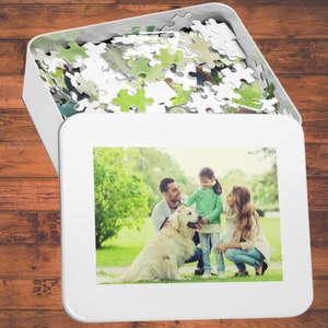 Peut inclure: Une famille de trois personnes, dont un chien, pose pour une photo dans un parc herbeux. La famille sourit et le chien regarde l'appareil photo. La photo est imprim&eacute;e sur une bo&icirc;te de puzzle blanche.