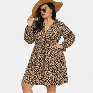 May include: A tan and black leopard print dress with long sleeves and a V-neck. The dress has a cinched waist and a flared skirt. The person in the image is wearing a wide-brimmed straw hat and sunglasses.