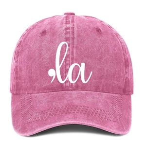 Puede incluir: Una gorra de béisbol de denim rosa con la palabra ".la" en letras blancas cursivas.