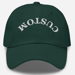 Puede incluir: Gorra de béisbol verde oscuro con la palabra "CUSTOM" bordada en blanco, arqueada en la parte delantera. La gorra tiene una visera curva y un cierre de correa de tela.