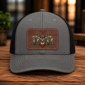 Puede incluir: Gorra de camionero gris con la parte trasera de malla negra. La parte delantera presenta un parche de cuero marrón con la palabra "DAD" en diseño de camuflaje y un gráfico de ciervo con gafas de sol y bandana.