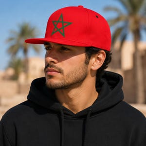 May include: A red baseball cap with a flat brim, featuring a green star emblem. The cap is worn by a person wearing a black hooded sweatshirt. The background includes palm trees and a building.